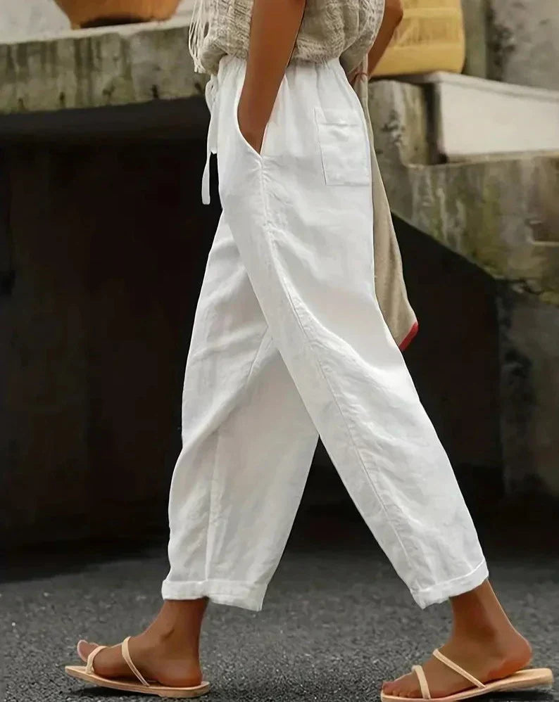 Personne portant un pantalon blanc ample en lin, style décontracté, avec des sandales beiges, marchant sur un trottoir urbain. Mode estivale chic.