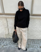 Femme en pull noir, pantalon beige ample, lunettes de soleil, sac noir, posant contre un mur. Mode automne, style décontracté chic.