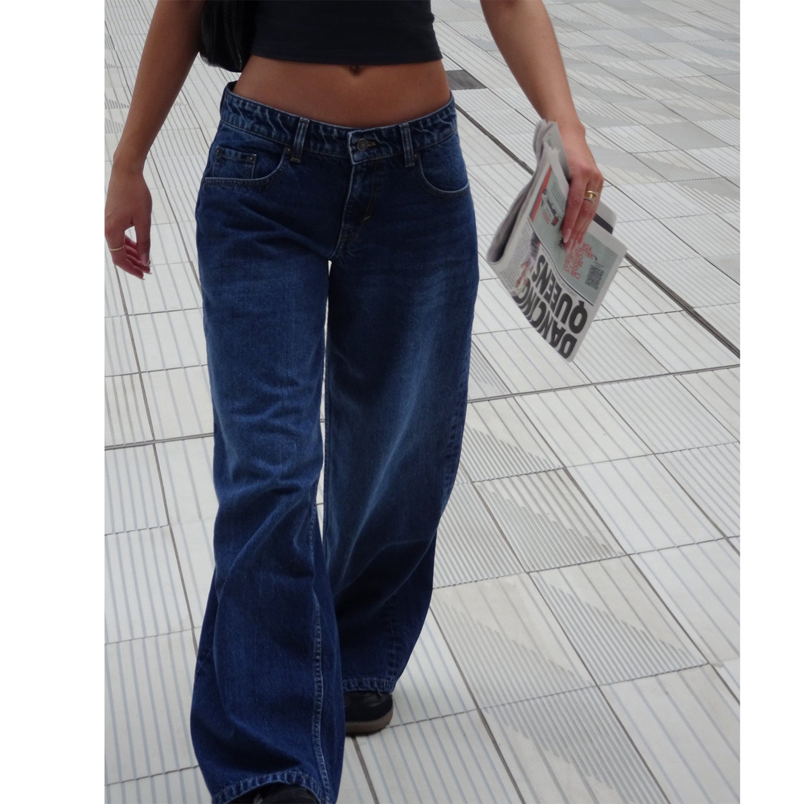 Jeune femme en jean large bleu, tenant un journal, marchant sur un trottoir moderne. Mode urbaine, style décontracté, tendance actuelle.