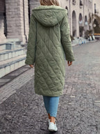Femme portant un manteau long matelassé vert avec capuche, marchant sur un trottoir pavé. Mode hiver, style décontracté, vêtements chauds.