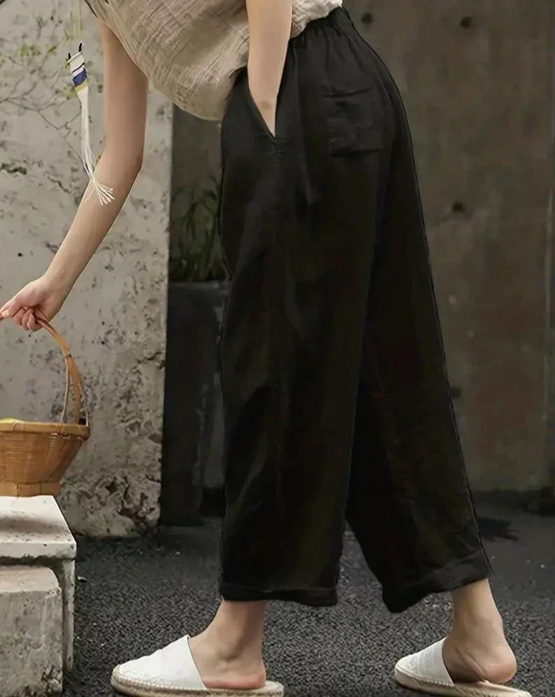 Pantalon large noir en lin pour femme, style décontracté, avec poches, porté avec des mules blanches. Mode estivale confortable et élégante.