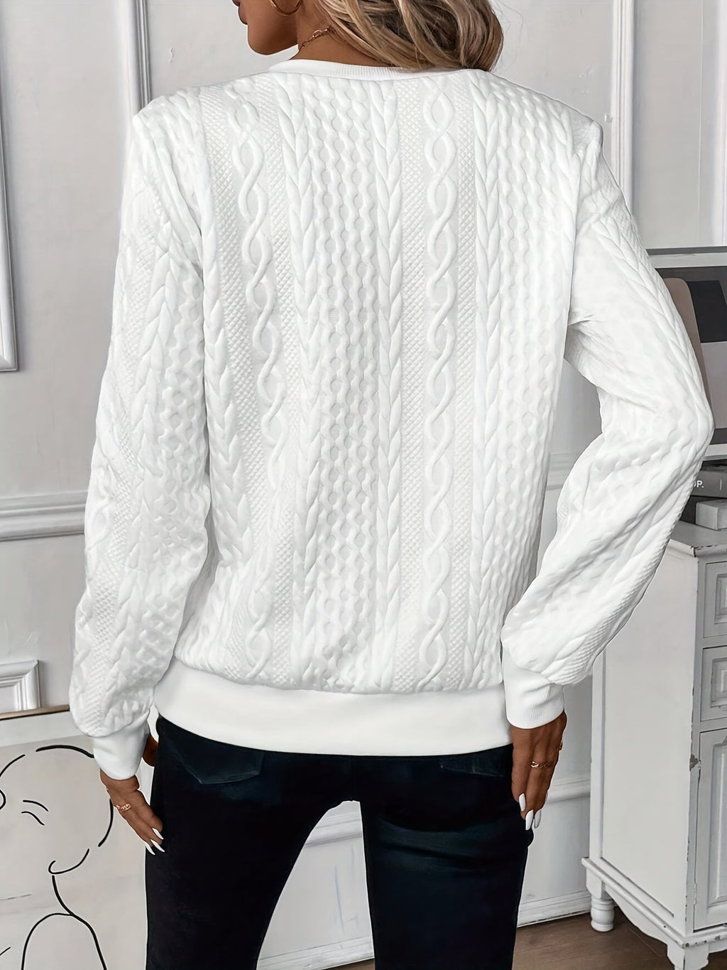 Femme portant un pull blanc en tricot torsadé, manches longues, dans un intérieur moderne. Mode automne-hiver, style élégant et confortable.