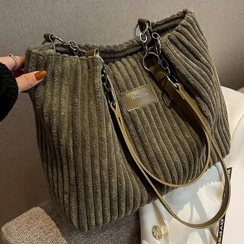 Sac à main en velours côtelé vert olive avec anses en cuir et chaîne, étiquette "Mollnuss", posé sur un canapé beige. Accessoire mode tendance.