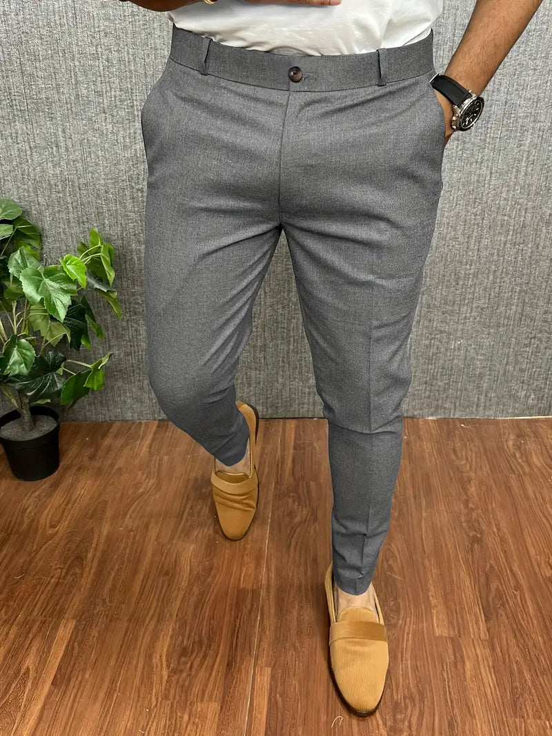 Pantalon gris élégant pour homme, porté avec des mocassins beiges et une montre noire, sur fond de sol en bois et plante verte. Mode masculine chic.