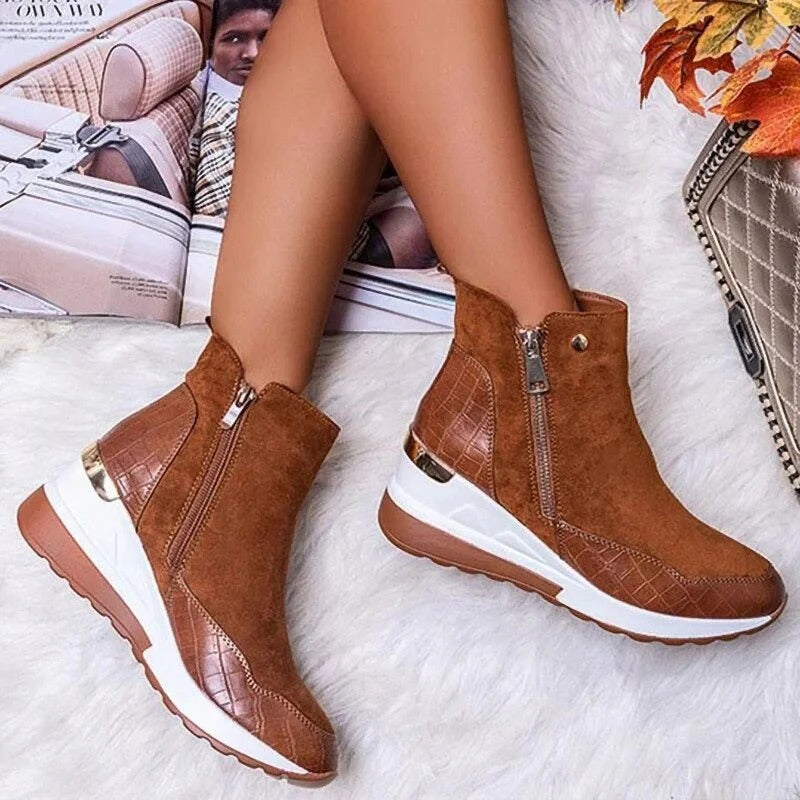 Bottes marron en daim avec fermeture éclair, semelle blanche et détails en cuir croco, portées par une personne assise sur un canapé beige.