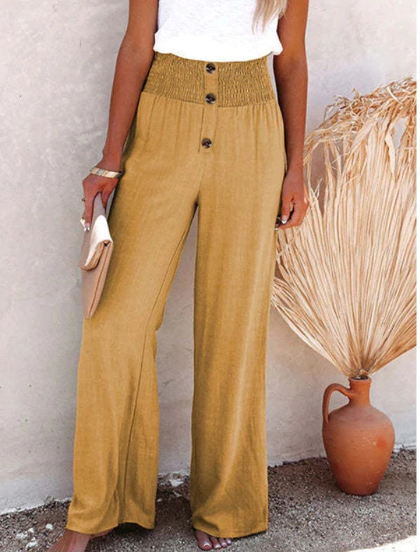 Femme portant un pantalon large jaune moutarde avec taille haute et boutons, tenant une pochette beige. Décor minimaliste avec vase en terre cuite.