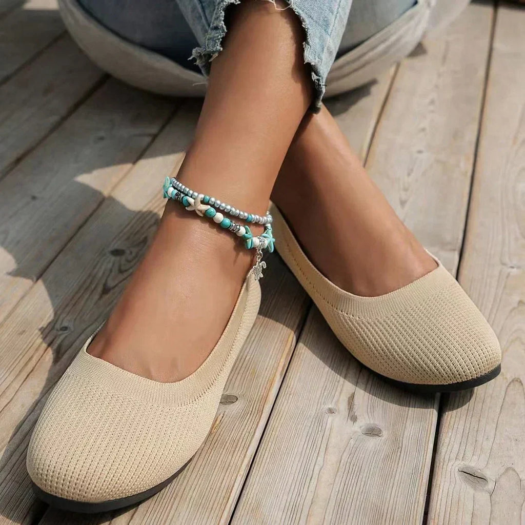 Ballerines beiges en tissu texturé sur pieds féminins, accompagnées d'un bracelet de cheville turquoise, posées sur un sol en bois.