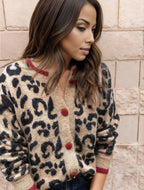 Femme portant un cardigan léopard beige et noir avec boutons rouges, posant devant un mur en pierre. Mode automne, style chic et tendance.