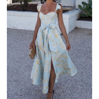 Robe d'été bleue à motifs floraux jaunes, avec ceinture nouée, portée par une femme tenant une pochette beige, sur fond extérieur ensoleillé.