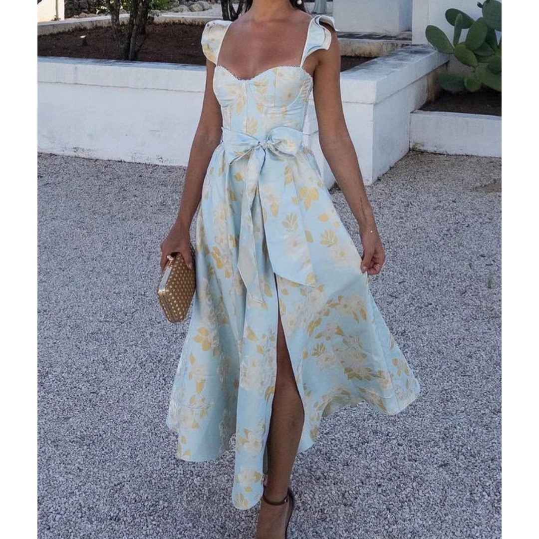 Robe d'été bleue à motifs floraux jaunes, avec ceinture nouée, portée par une femme tenant une pochette beige, sur fond extérieur ensoleillé.