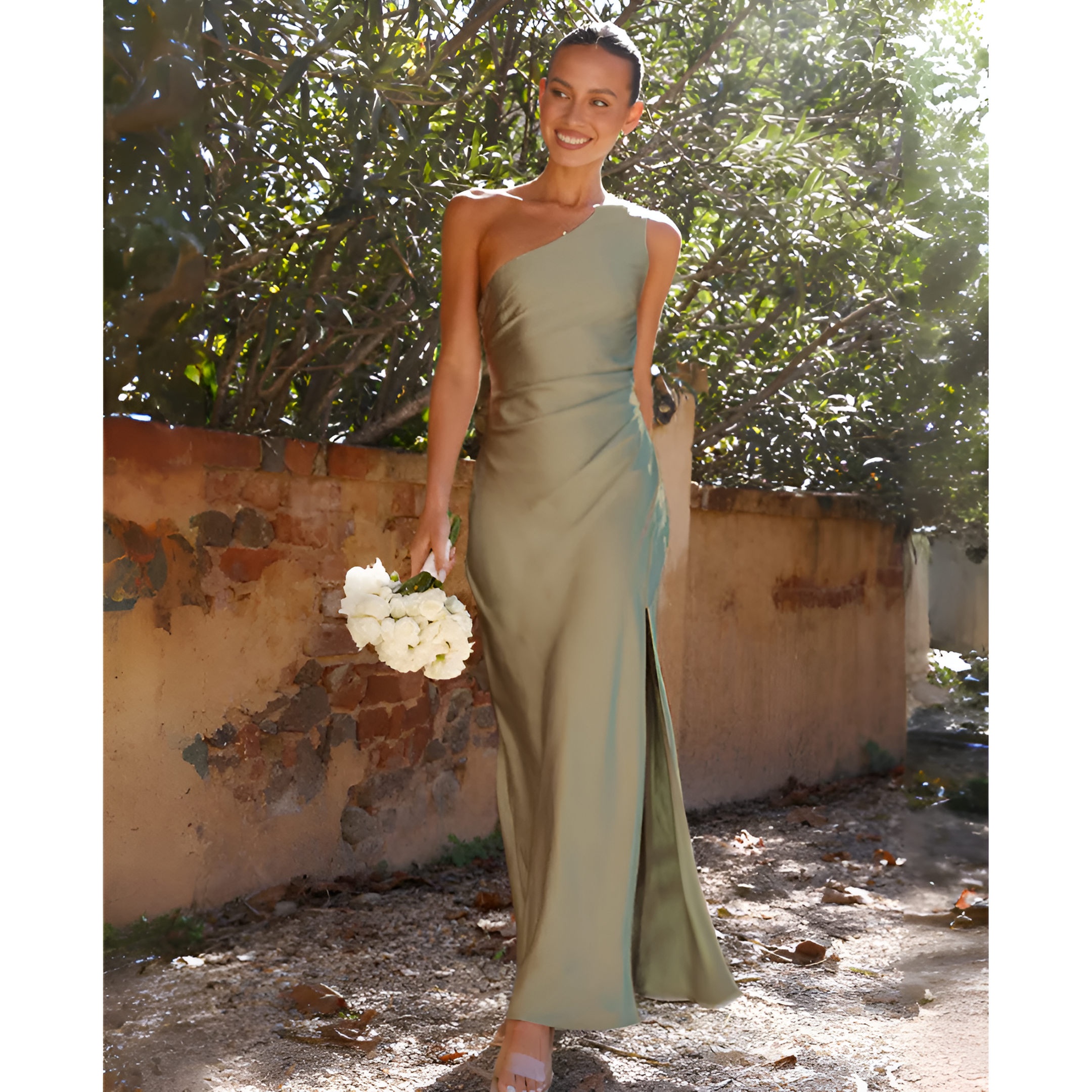 Une femme souriante en robe de soirée verte asymétrique, tenant un bouquet de fleurs blanches, marche dans un jardin ensoleillé.