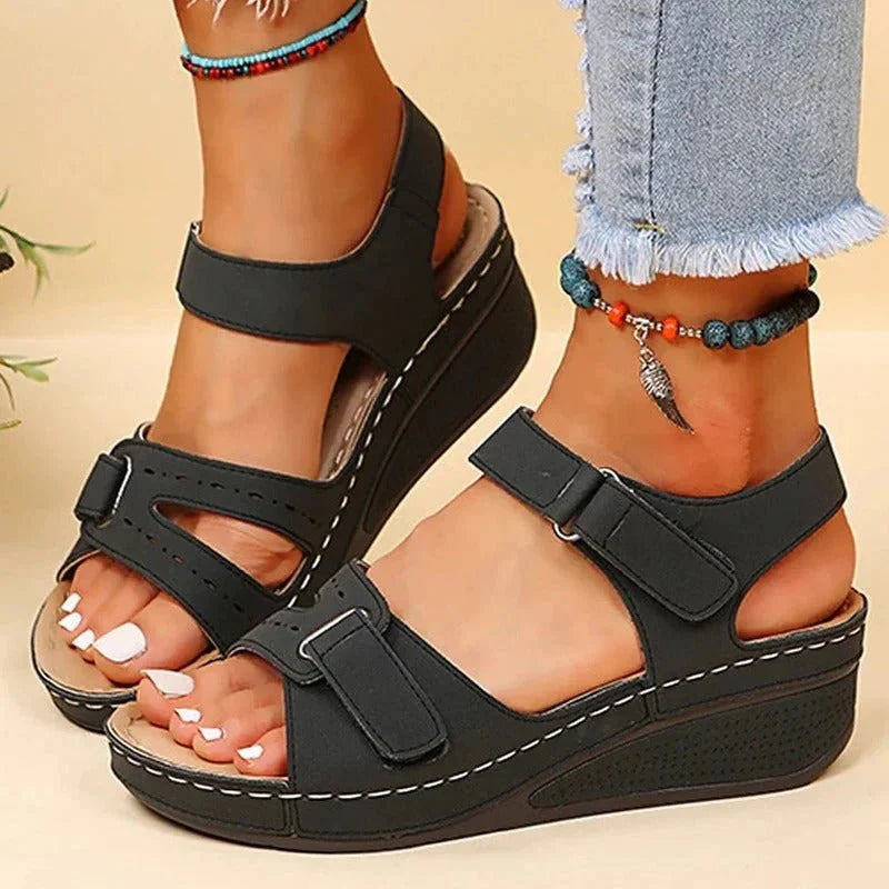 Sandales compensées noires pour femme avec brides réglables, portées avec un jean décontracté et un bracelet de cheville. Mode estivale tendance.