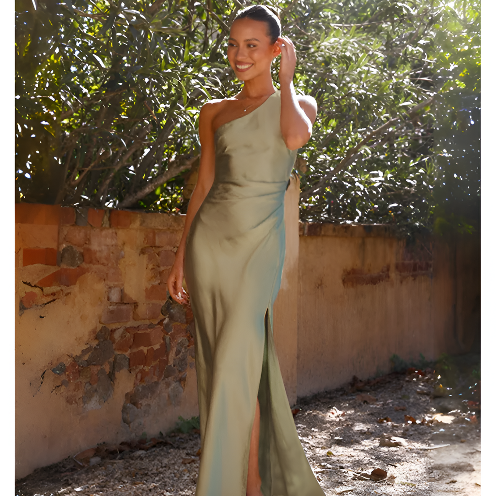 Robe de soirée verte élégante, asymétrique, avec fente, portée par une femme souriante dans un jardin ensoleillé, mur de briques en arrière-plan.