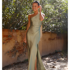 Robe de soirée verte élégante, asymétrique, avec fente, portée par une femme souriante dans un jardin ensoleillé, mur de briques en arrière-plan.