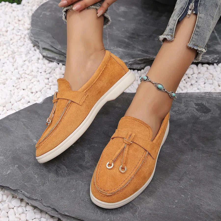 Mocassins en daim orange pour femme, élégants et confortables, portés avec un jean déchiré. Chaussures tendance sur fond de galets blancs.