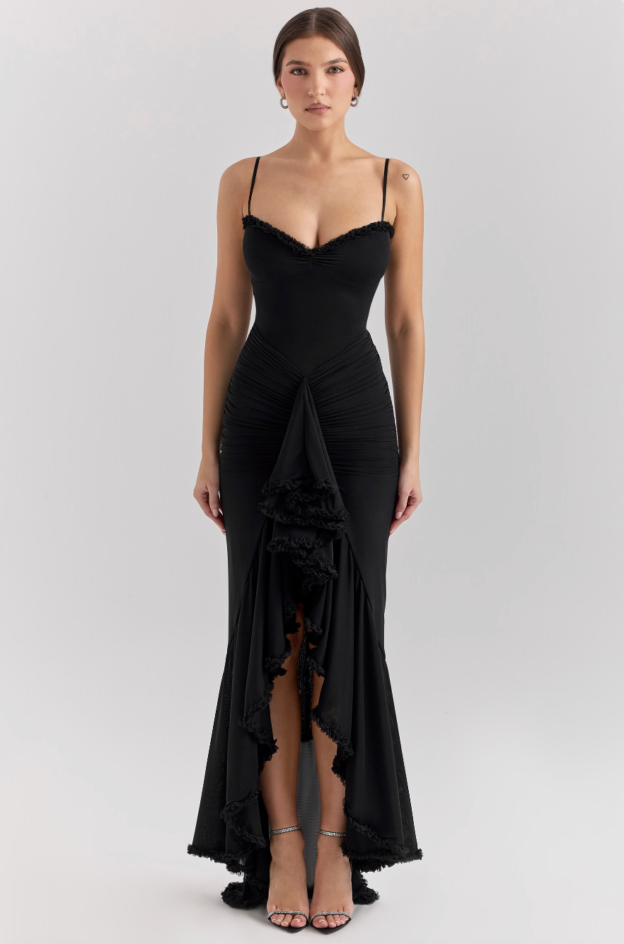 Femme portant une élégante robe noire longue avec bretelles fines et détails en dentelle, posant debout. Mode soirée, style glamour.