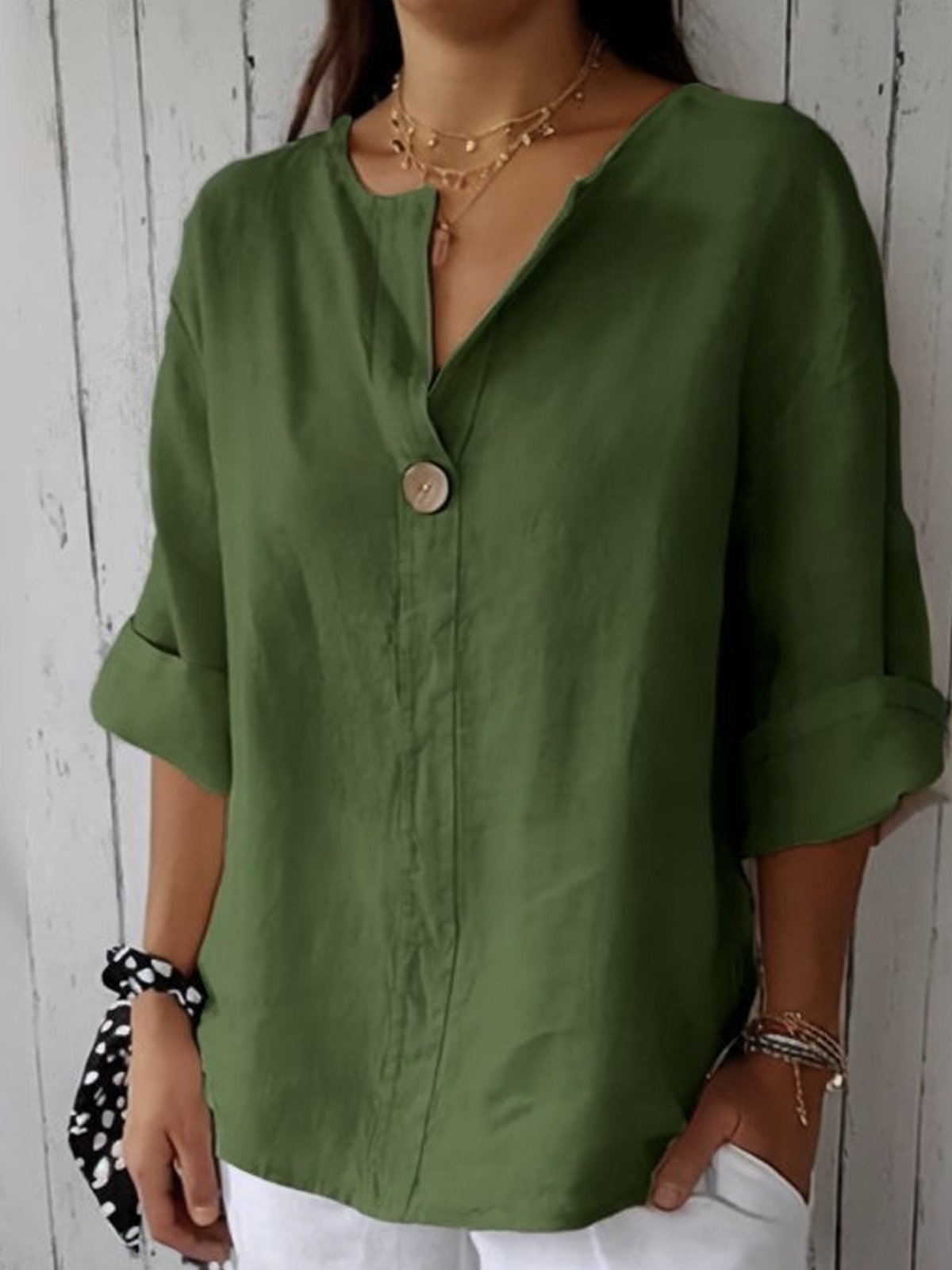 Femme portant une chemise verte en lin avec manches retroussées, bouton en bois, collier doré, sur fond de mur en bois blanc. Mode décontractée élégante.