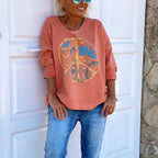 Femme souriante portant un sweat-shirt rose avec motif peace and love, lunettes de soleil, devant une porte blanche. Mode décontractée, style bohème.