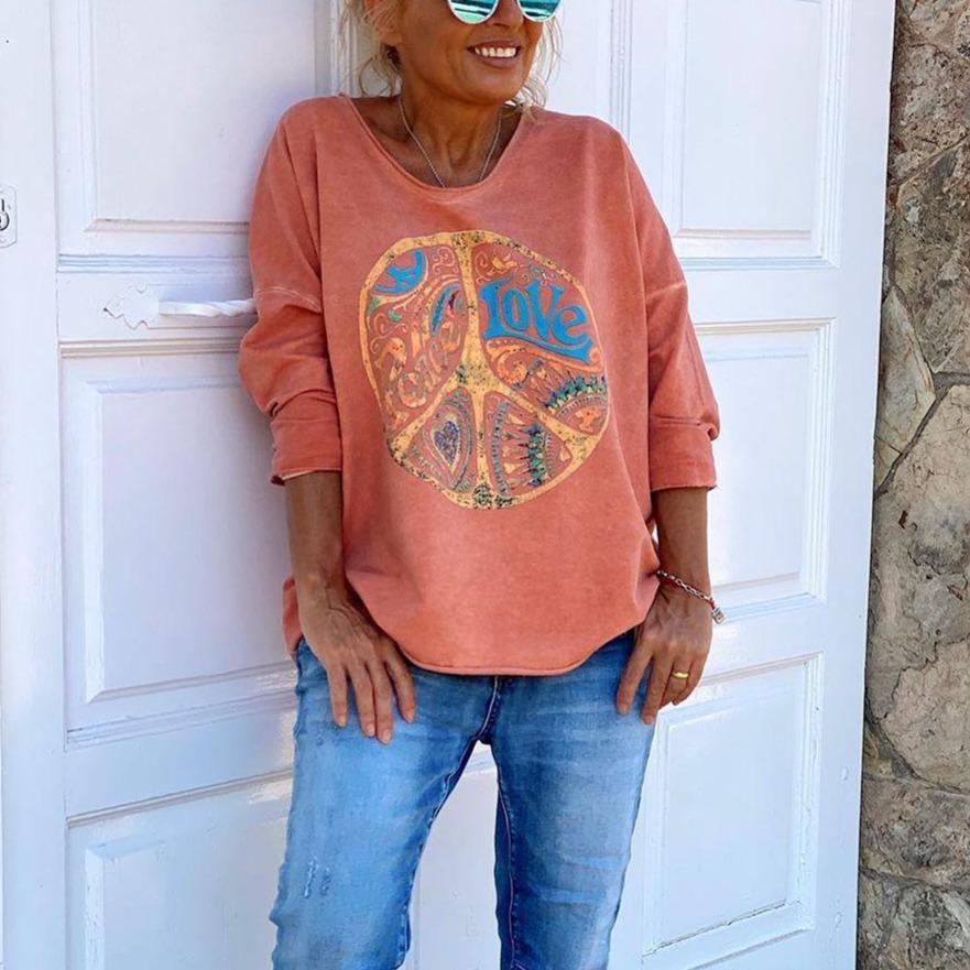 Femme souriante portant un sweat-shirt rose avec motif peace and love, lunettes de soleil, devant une porte blanche. Mode décontractée, style bohème.
