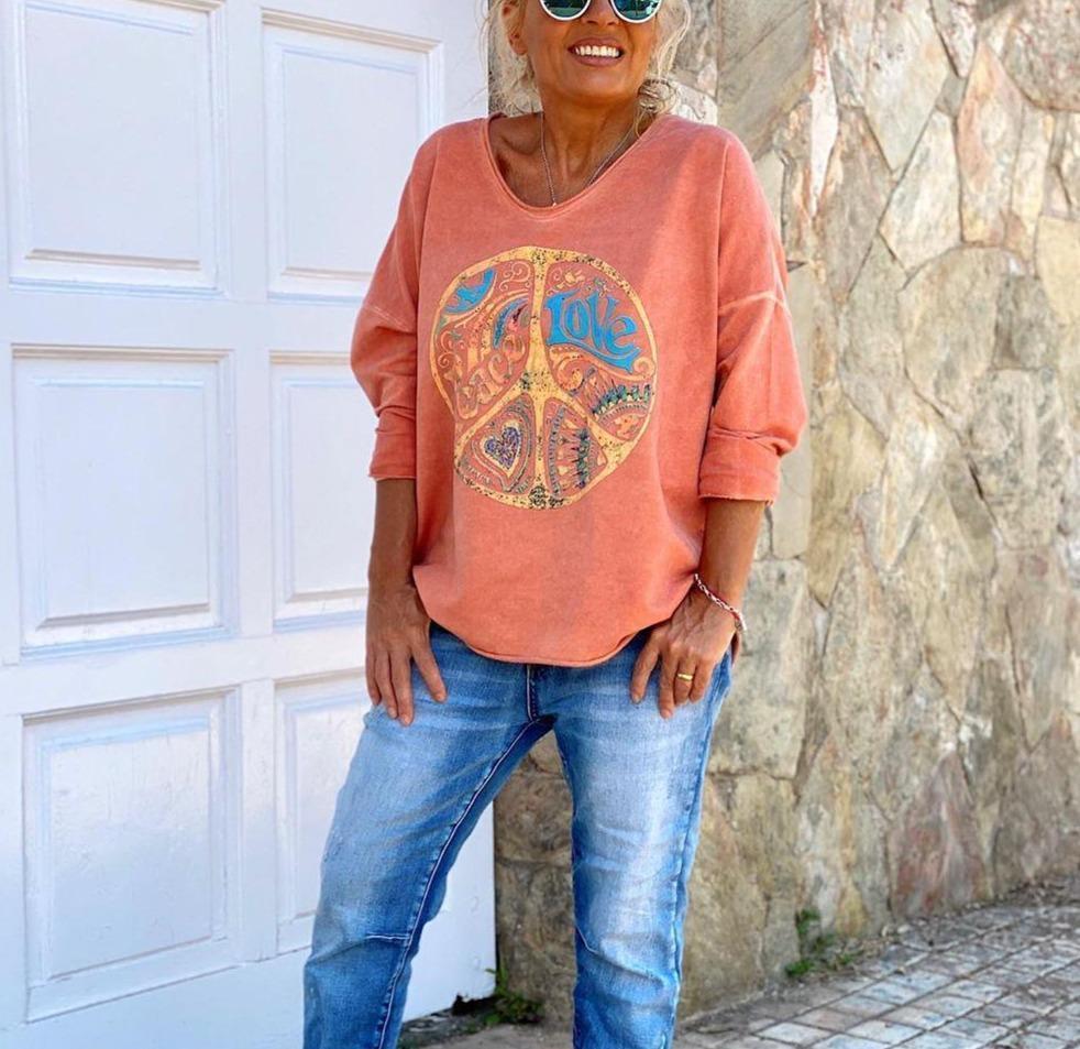 Femme souriante portant un pull orange avec symbole peace and love, lunettes de soleil, jeans décontractés, devant une porte blanche et mur en pierre.