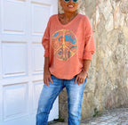 Femme souriante portant un pull orange avec symbole peace and love, lunettes de soleil, jeans décontractés, devant une porte blanche et mur en pierre.