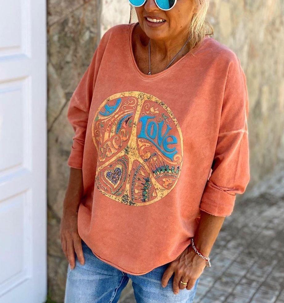 Femme portant un sweat-shirt orange avec symbole peace and love coloré, lunettes de soleil bleues, devant une porte blanche. Mode décontractée, style bohème.