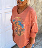Femme souriante portant un pull orange avec motif peace and love, lunettes de soleil bleues, devant une porte blanche. Mode décontractée, style bohème.