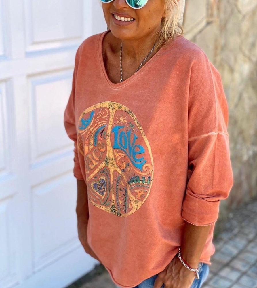 Femme souriante portant un pull orange avec motif peace and love, lunettes de soleil bleues, devant une porte blanche. Mode décontractée, style bohème.