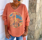 Femme souriante portant un pull orange avec motif Peace and Love, lunettes de soleil bleues, posant devant un mur en pierre. Mode décontractée.
