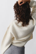 Femme de dos portant un pull en laine blanc et une jupe grise, cheveux bouclés. Mode automne-hiver, style décontracté et élégant.