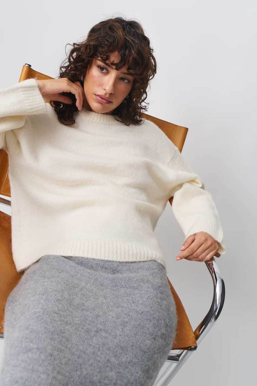 Femme assise sur une chaise en cuir, portant un pull blanc en laine et une jupe grise. Mode automne-hiver, style décontracté et élégant.