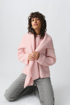 Femme aux cheveux bouclés portant un pull rose et un châle, posant sur fond blanc. Mode féminine, style décontracté, vêtements d'hiver.