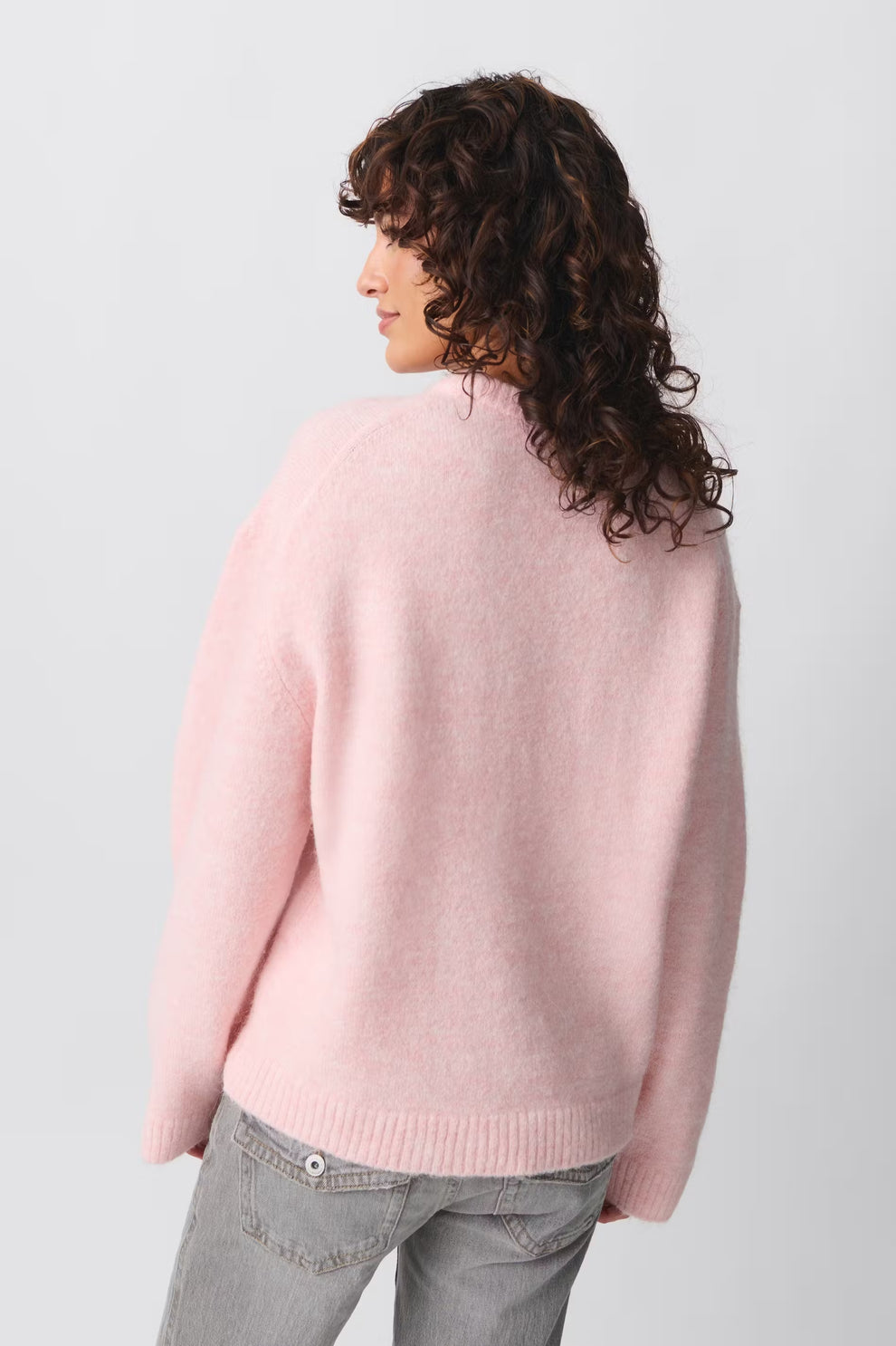 Femme portant un pull rose en laine, vue de dos, cheveux bouclés, jeans gris. Mode automne-hiver, style décontracté, tendance féminine.