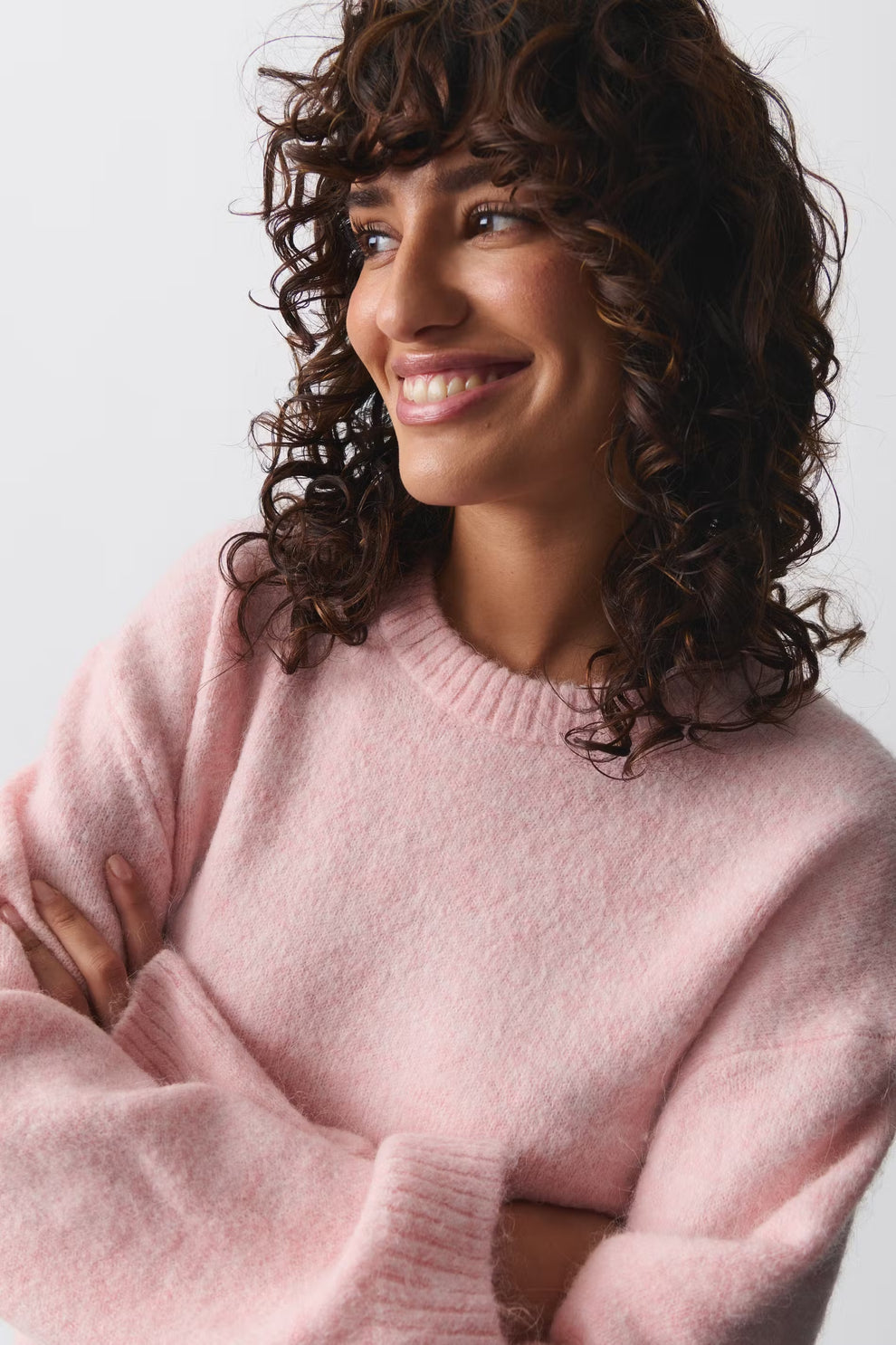 Femme souriante portant un pull rose en laine, bras croisés, cheveux bouclés. Mode féminine, style décontracté, tendance automne-hiver.