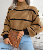 Femme portant un pull en maille marron à rayures noires, tenant un sac à main en cuir marron à franges, assise sur une commode blanche.