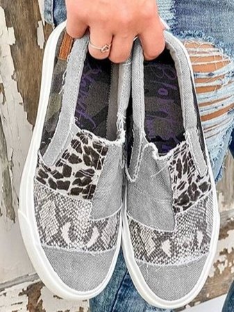 Chaussures slip-on en toile grise avec motifs léopard et serpent, tenues par une main. Style décontracté, mode urbaine, jeans déchiré en arrière-plan.