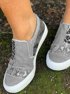 Chaussures slip-on grises à motifs camouflage et léopard, semelle blanche, portées sur sol moussu. Sneakers tendance pour femme, style décontracté.