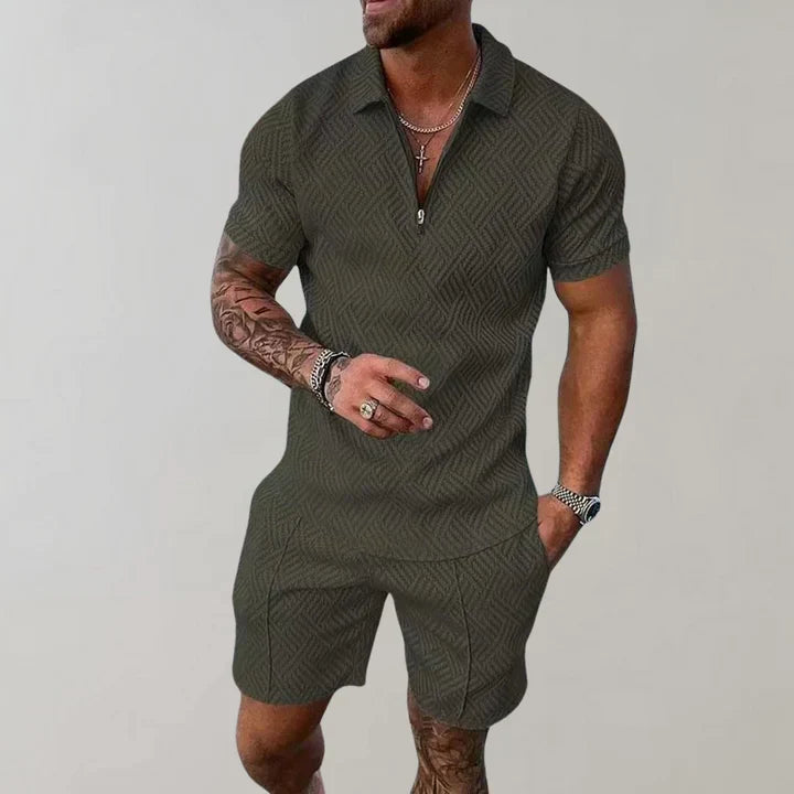 Homme en tenue décontractée, combinaison verte à motifs géométriques, manches courtes, col zippé, bras tatoués, accessoires argentés.