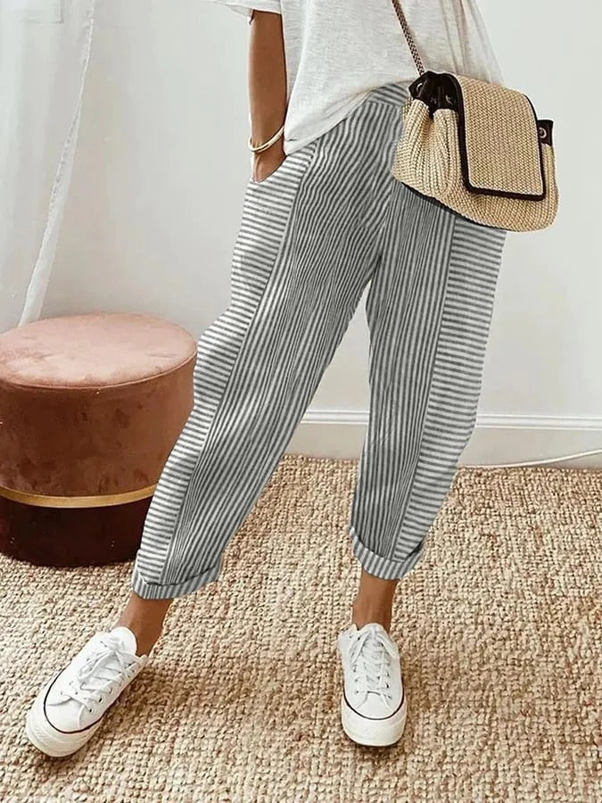 Femme portant un pantalon rayé gris et blanc, t-shirt blanc, baskets blanches, sac en osier. Mode décontractée, tendance estivale, style chic.