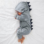 Bébé endormi en pyjama dinosaure gris avec capuche et écailles noires, allongé sur un lit blanc. Pyjama bébé mignon, sommeil paisible.