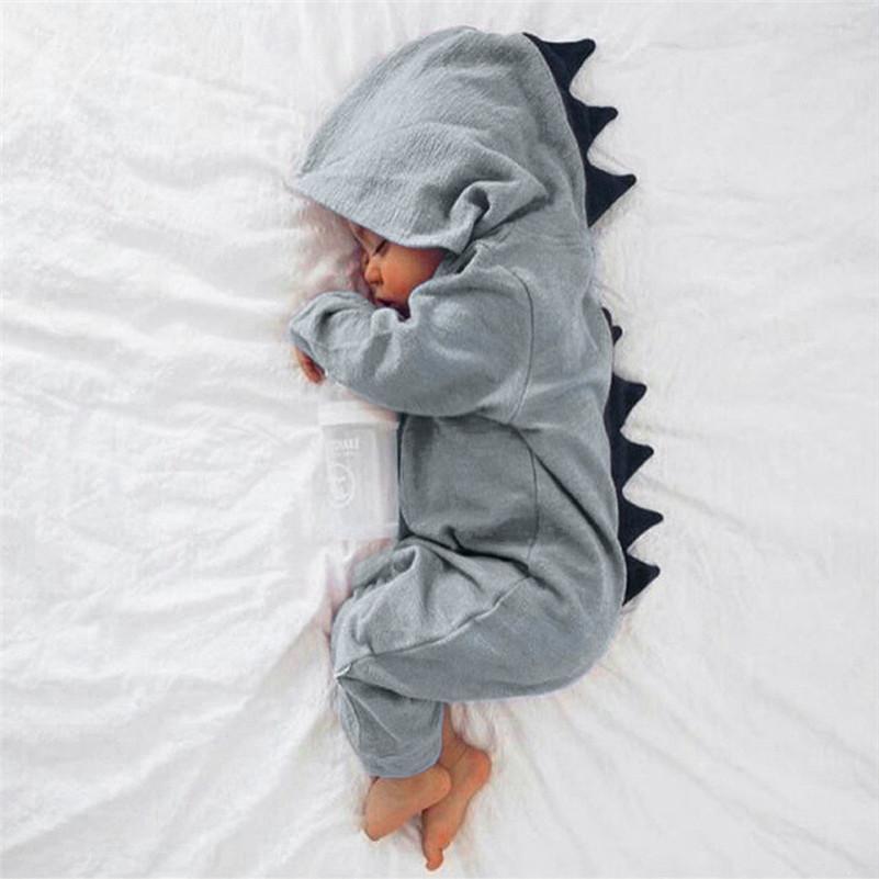 Bébé endormi en pyjama dinosaure gris avec capuche et écailles noires, allongé sur un lit blanc. Pyjama bébé mignon, sommeil paisible.