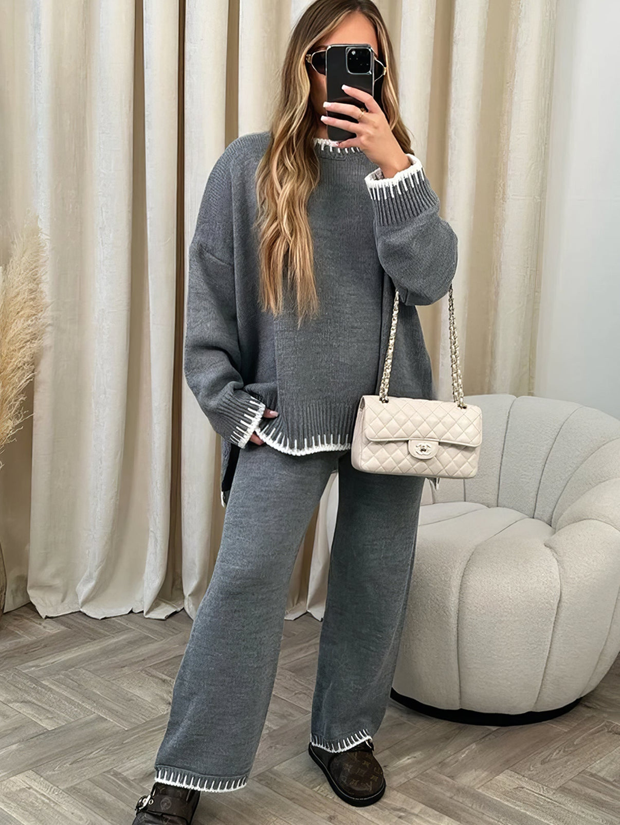 Femme en ensemble pull et pantalon gris, sac à main blanc, posant devant un miroir. Mode automne-hiver, tenue décontractée élégante.