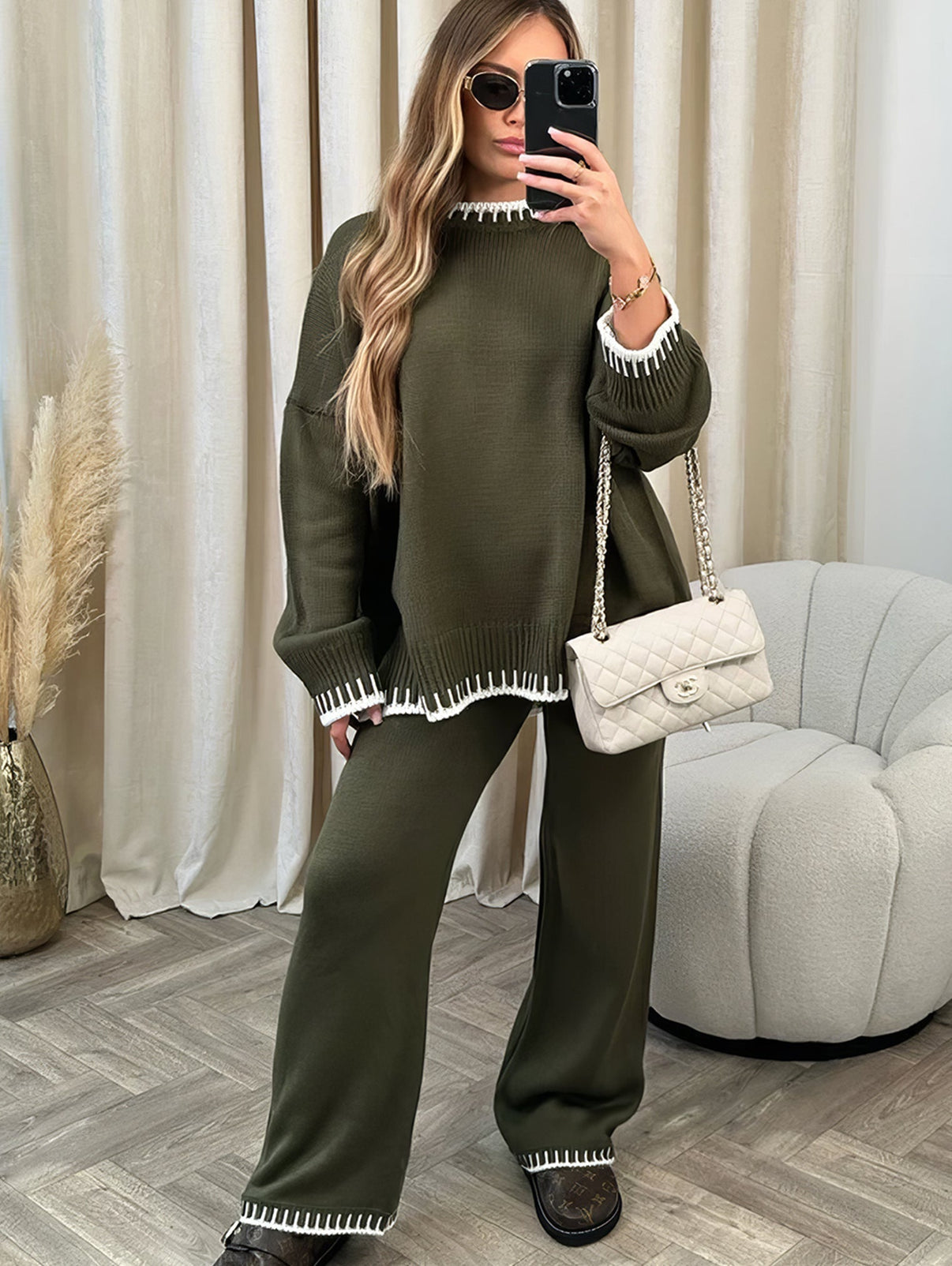 Femme en ensemble vert olive chic, pull et pantalon, sac à main blanc, lunettes de soleil, intérieur moderne. Mode automne-hiver tendance.