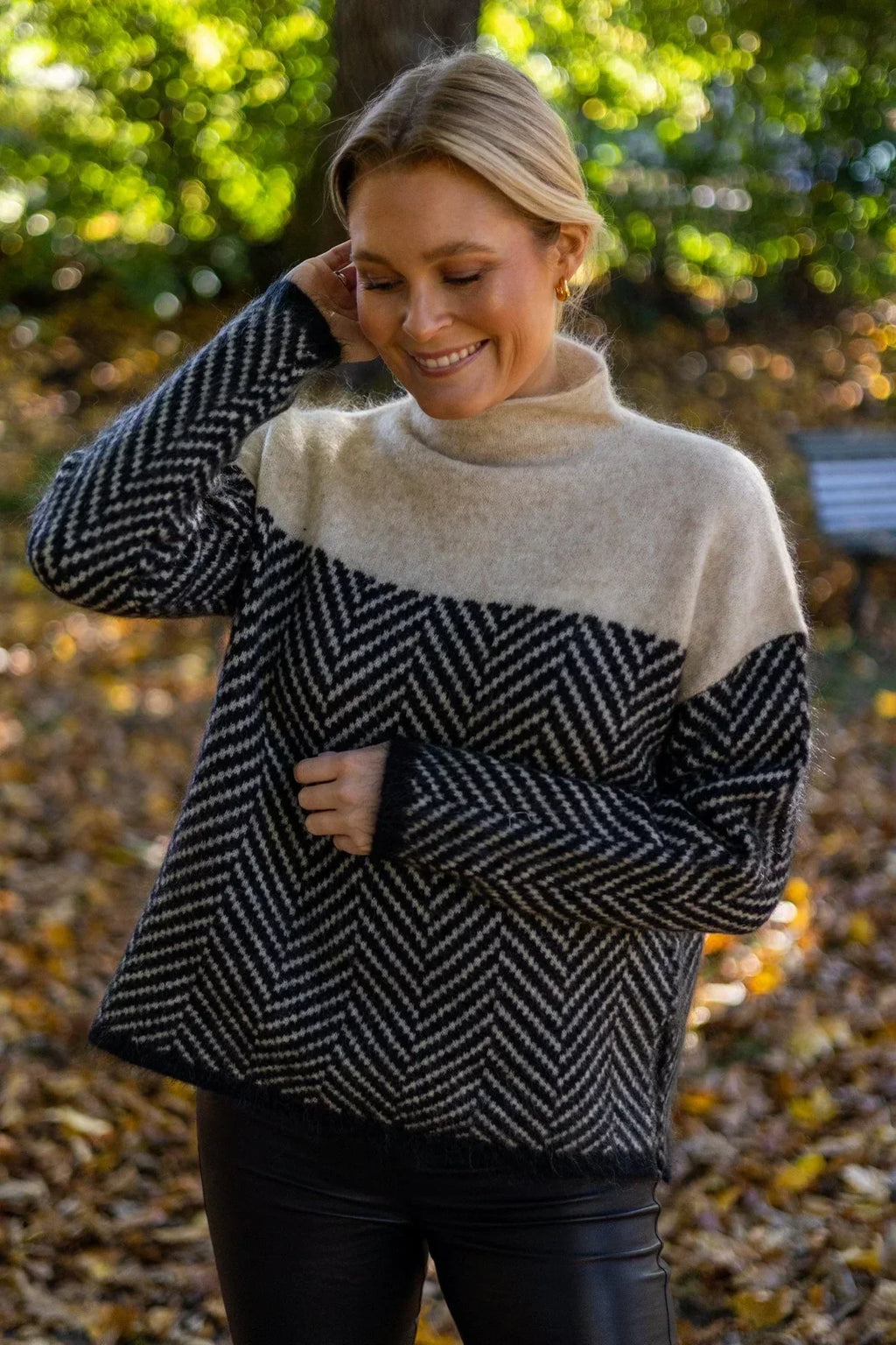 Femme souriante portant un pull à motifs géométriques noir et blanc, col roulé, dans un parc automnal. Mode automne, style décontracté, nature.