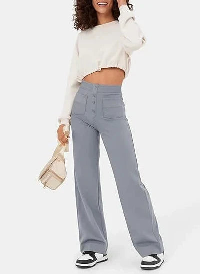 Femme souriante portant un pull blanc court et un pantalon large gris taille haute, tenant un petit sac beige. Mode décontractée tendance.