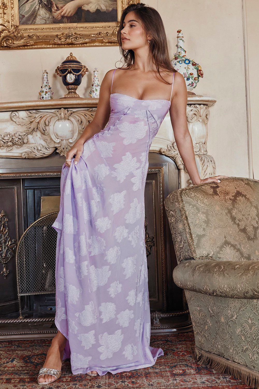 Femme en robe longue violette à motifs floraux, posant élégamment dans un salon vintage avec cheminée décorative et fauteuil ancien.