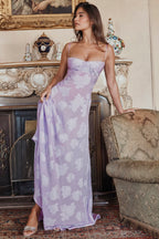 Femme en robe longue violette à motifs floraux, posant élégamment dans un salon vintage avec cheminée décorative et fauteuil ancien.