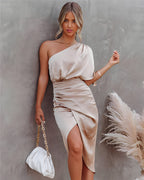 Femme en robe satinée beige asymétrique, tenant un sac blanc à chaîne dorée, posant contre un mur gris avec des herbes de pampa en arrière-plan.