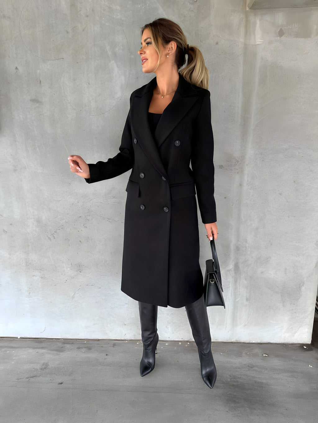 Femme élégante en manteau noir, bottes hautes et sac à main, posant contre un mur gris. Mode automne-hiver, style chic et moderne.