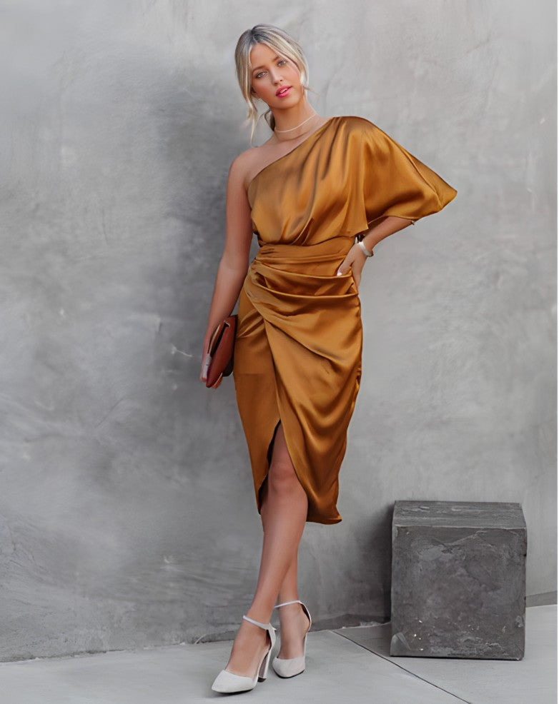 Femme en robe asymétrique dorée, posant contre un mur gris, tenant une pochette marron, portant des talons blancs. Mode élégante et moderne.